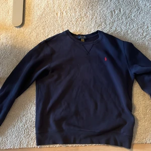 Marinblå sweatshirt från Polo Ralph Lauren - Snygg marinblå sweatshirt från Polo Ralph Lauren med klassisk röd broderad logga på bröstet. Tröjan har rund hals, långa ärmar och är tillverkad i mjuk bomull. Perfekt för dig som gillar stilrena och tidlösa plagg med en sportig touch.