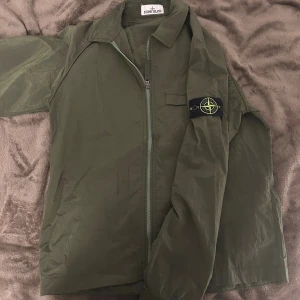 Stone Island grön overshirt jacka - Snygg militärgrön overshirt jacka från Stone Island med klassisk kompasspatch på vänster ärm. Jackan har dragkedja framtill, krage och är tillverkad i lätt polyester. Perfekt för dig som vill ha en clean och stilren look med street-vibe.