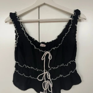 Svart volangtopp med snörning från Nelly - Supersöt svart topp från Nelly med vita kontrastsömmar och volangdetaljer. Strl M. 