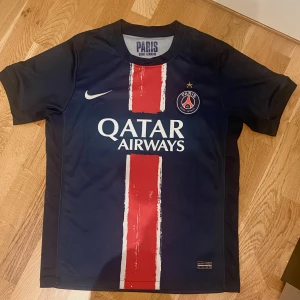 PSG Nike Dri-Fit  - Paris Saint-Germain fotbollströja från Nike i Dri-Fit-material. Mörkblå med en bred röd rand och vita detaljer, PSG-emblem på bröstet och Qatar Airways-tryck. Kortärmad och tillverkad i Thailand. Perfekt för dig som älskar fotboll och vill bära klubbens färger.