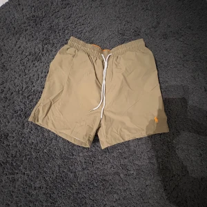 Khaki badshorts från Polo Ralph Lauren - Snygga khaki badshorts från Polo Ralph Lauren med vit snörning i midjan och orange insida. Klassisk design med elastisk midja, bakficka med lock och meshfoder. Perfekta för stranden eller poolen. Pris kan diskuteras, hör av dig vid minsta fråga!