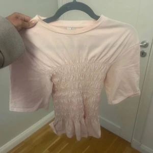 Ljusrosa smockad topp från & Other Stories - Supersöt ljusrosa topp från & Other Stories i bomull. Modellen har korta ärmar, smockad midja och lätt volang nertill som ger en feminin vibe. Perfekt till jeans eller kjol för en chill och söt look. Buda gärna ❤️