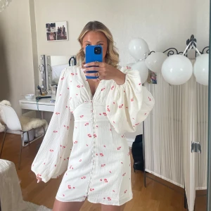 Vit kortklänning med blommigt mönster - Supersöt vit kortklänning med små röda blommor, v-ringning och en figurnära passform över midjan. Perfekt för sommarens alla tillfällen! Den populära modellen från adoore. Använd 1 gång, ser ut exakt som ny. 