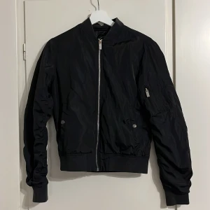 Svart bomberjacka från Crocker XS - Svart bomberjacka från Crocker i storlek XS. Jackan har klassisk rak passform, dragkedja framtill, fickor med tryckknapp och en ärmficka med dragkedja. Tillverkad i 100% polyester med ribbade muddar vid ärmslut och midja.