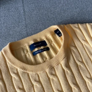 Ljusgul kabelstickad tröja GANT - Snygg ljusgul kabelstickad tröja från GANT i stretchig bomull. Klassisk rund hals och diskret broderad logga på bröstet. Perfekt för dig som gillar preppy stil och vill ha en mjuk, bekväm tröja med lite extra färg.