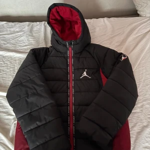 Svart och röd Jordan pufferjacka/vinter jacka - Säljer en snygg pufferjacka från Jordan i svart och rött med ikonisk Jumpman-logga på bröstet och Air-märke på ärmen. Jackan har huva med röd insida, dragkedja med Jordan-tryck och quiltad design. Perfekt för dig som vill ha streetstyle och värme. 100% bomull 12-13 åringar 147-163cm
