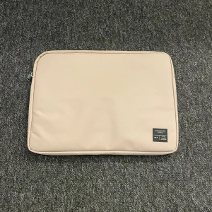 Beige datorväska från 157 Denim Makers - Stilren beige datorväska från 157 Denim Makers med svart patch framtill. Väskan är rektangulär och har dragkedja längs kanten. Tillverkad i slitstark polyester som skyddar din laptop på ett snyggt sätt.