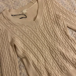Beige kabelstickad tröja H&M - Kabelstickad tröja från H&M i beige med v-ringning och långa ärmar. Snyggt flätmönster över hela tröjan och bred ribbad kant vid halsen. Perfekt för lager-på-lager under hösten och vintern.