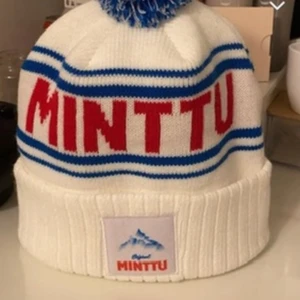Minttu vit mössa med blå tofs - Säljer en vit stickad Minttu-mössa med blå och röda detaljer. Mössan har bred ribbad kant, blå ränder och stor blå-vit tofs på toppen. Framtill finns ett Minttu-märke med bergmotiv. Perfekt för kalla dagar och snygg streetstil. Helt oanvänd säljer då jag fick två stycken i present men behöver ba en, pris kan absolut diskuteras!