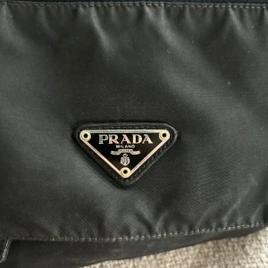 Vintage Prada Tessuto Messengerbag i Nylån - Säljer en vintage axelväska från Prada med klassisk trekantig logga framtill. Väskan är tillverkad i slitstark nylon med detaljer i skinn och har justerbar axelrem, dragkedja och spännen. Perfekt för dig som vill ha en stilren och praktisk väska med lyxig känsla. Köpt från The Real Real, kvitto finns.