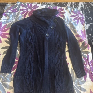 Mörkblå stickad kofta med knappar - Säljer en mörkblå lång kofta med grovstickade flätor längs framsidan och stora knappar i sned knäppning. Koftan har höga ribbade muddar, två fickor framtill och en mysig, hög krage. Perfekt för kyliga dagar och enkel att styla till jeans eller leggings.
