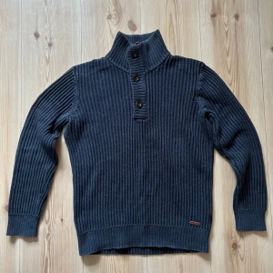 Mörkblå stickad tröja Peak Performance - Mörkblå ribbstickad tröja från Peak Performance med hög krage och tre knappar framtill. Tröjan har lång ärm och är tillverkad i ett mjukt, stickat material. Perfekt för kyliga dagar och snygg till jeans eller cargobyxor.