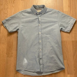 Ljusblå kortärmad skjorta Tailored Originals - Ljusblå skjorta från Tailored Originals med korta ärmar och button-down krage. Skjortan har klassisk passform, vita knappar och är tillverkad i bomull. Perfekt för en clean och avslappnad stil.