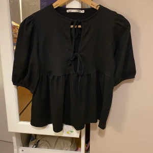 Svart blus med knyt från 157 - Svart blus från 157 i storlek XS med puffiga halvlånga ärmar och två knytband framtill. Blusen har en lätt rynkad midja och är tillverkad i ett strukturerat tyg som ger en cool känsla. Perfekt för dig som gillar en avslappnad men ändå trendig stil.