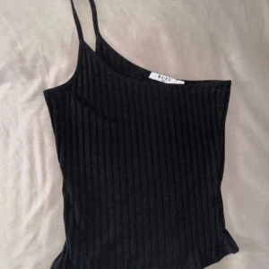 Svart ribbad linnetopp från NA-KD - Säljer en svart ribbad linnetopp från NA-KD med asymmetrisk one shoulder-design och smala axelband. Toppen är tight och har en stilren look som passar perfekt till jeans eller kjol. Materialet är mjukt och stretchigt för en bekväm känsla. Toppen är lite nopprig på sidorna men annars i fint skick!