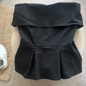 Svart axelbandslös tubtopp med volang - Säljer en svart axelbandslös tubtopp med bred volang upptill och utsvängd nederdel. Mycket bra skick😊