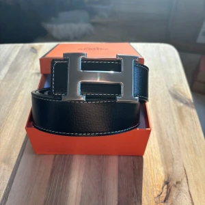 Svart bälte med silver spänne - Säljer ett svart bälte. Bältet är gjort i exklusivt skinn med vit kontrastsöm och levereras i original orange box. Perfekt accessoar för att lyfta vilken outfit som helst.