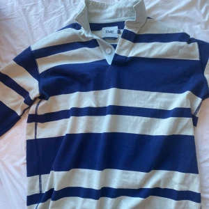 Randig pikétröja från Drake's i blått - Säljer en klassisk pikétröja från Drake's i storlek M. Tröjan har breda marinblå och beige ränder, lång ärm och krage med två knappar. Tillverkad i mjuk bomull, perfekt för dig som gillar sportig och chill stil. Nypris ca 3500kr