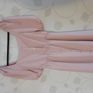 Ljusrosa klänning med puffärmar - Supersöt ljusrosa klänning med puffiga kortärmar och knappar framtill. Klänningen har en markerad midja och en rak, enkel siluett. Perfekt för dig som gillar en feminin och trendig look. Materialet känns lätt och mjukt mot huden.