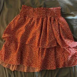 Röd blommig volangkjol  - Supersöt röd kortkjol med vita och gula blommor, bred smock i midjan och dubbla volanger. Det finns ett litet hål där den ander volangen sitter men det är inget man ser när man har på sig den. 