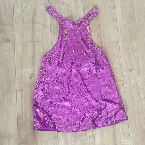 Glittrig lila paljettklänning - Säljer en kort lila klänning täckt med glittriga paljetter i guld och lila. Klänningen har en ärmlös design med breda axelband och rundad hals. Materialet är syntet med en mjuk insida och hela framsidan är täckt av paljetter🩷💜