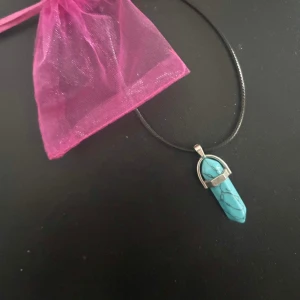 Halsband med turkos kristallhänge - Snyggt halsband med ett turkost kristallformat hänge och silverfärgad infattning. Hängs i ett svart snöre och levereras i en rosa organzapåse. Perfekt accessoar för att lyfta din outfit.