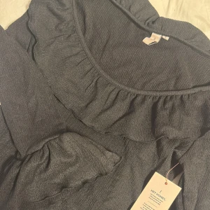 Svart volangblus från Nelly - Supersöt svart blus från Nelly med volangdetaljer längs ringningen och vid ärmsluten. Blusen har lång ärm och är tillverkad i ett lätt, strukturerat tyg som ger en snygg passform. Aldrig använd, prislappen är kvar. 