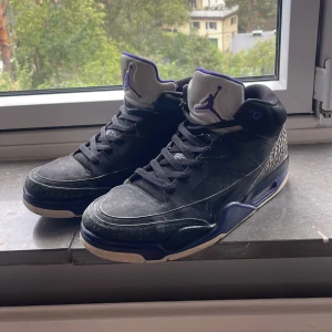 Nike Air Jordan sneakers svart/lila - Säljer ett par Nike Air Jordan sneakers i svart med lila detaljer och klassiskt elephant print vid hälen. Skorna har snörning, rund tå och platt sula. Insidan är fodrad i lila textil och tungan pryds av den ikoniska Jumpman-loggan. Perfekta för dig som gillar streetwear och unika detaljer.