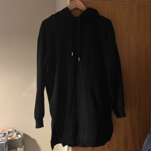 Svart hoodieklänning från H&M - Svart lång hoodie från H&M Basics med huva och dragsko. Modellen är oversized och har långa ärmar, ribbade muddar och slitsar i sidorna. Perfekt för chill dagar eller när du vill vara extra bekväm.
