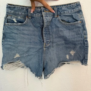 Blå jeansshorts med slitna detaljer från Zara - Snygga blå jeansshorts från Zara med hög midja och råa, fransiga kanter. Shortsen har slitna detaljer framtill och klassiska fem fickor. Perfekta för en avslappnad och trendig look i sommar.