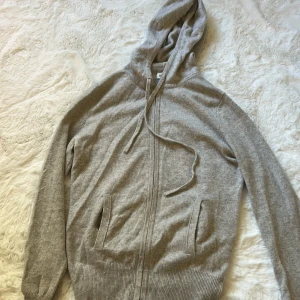 Grå hoodie i kashmir från Soft Goat - Mjuk och lyxig grå hoodie från Soft Goat i kashmir. Tröjan har huva med snörning, dragkedja framtill och två fickor. Långärmad med ribbade muddar vid ärmslut och nederkant. 