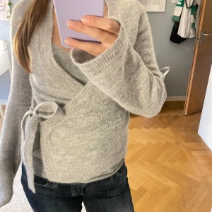 Grå omlottkofta från Gina Tricot - Mysig grå omlottkofta från Gina Tricot i ullblandning. Koftan har långa ärmar, v-ringad hals och knyt i sidan som ger en snygg passform. Perfekt att slänga på sig när det är lite kyligt ute. Stilren och enkel att matcha med jeans eller kjol.