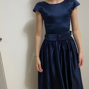 Marinblå långklänning med rosett - Elegant marinblå långklänning i glansigt tyg med markerad midja och bred rosett baktill. Klänningen har korta ärmar, rund halsringning och ett vackert fallande kjolparti. Perfekt för dig som vill sticka ut med en klassisk och stilren look.