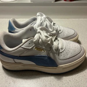 Vita och blå sneakers från Puma - Snygga sneakers från Puma i vitt och ljusblått med klassisk design. Skorna har snörning, perforerad tå och detaljer i skinn och syntet. Guldiga Puma-loggan på sidan och blå logga på plösen. Perfekta för dig som gillar stilrena och sportiga vibes. Använda fåtal gånger, som nya. 