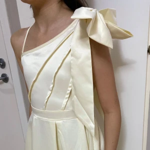 Champagnefärgad långklänning med rosett - Elegant långklänning i champagnefärgad satin med oneshoulder och stor rosett på axeln. Klänningen har veckad kjol och markerad midja, vilket ger en lyxig och stilren look. Perfekt för dig som vill sticka ut med en unik och sofistikerad stil.