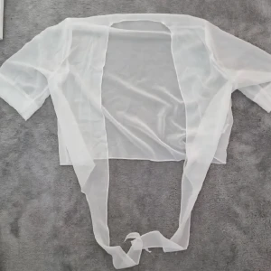 Vit transparent bolerotopp med knyt S-L - Säljer en superfin vit bolerotopp i transparent material. Toppen har korta ärmar och långa band framtill som kan knytas för en snygg look. Perfekt att styla över linne eller klänning för en trendig layering-effekt.
