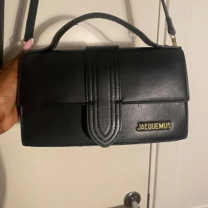 Svart axelväska från Jacquemus - Säljer en stilren svart axelväska från Jacquemus i slätt skinn med guldfärgad logga framtill. Den är väldigt identisk till den riktiga men skillnaden är bara att den inte har en ficka bakom. Har använt några gånger och mer bilder kan skickas💗🫶🏾 