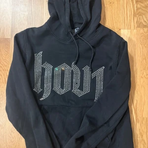 Hov1 merch  - Svart hoodie med huva och snörning, dekorerad med stora bokstäver i glittrande strass på bröstet. Mjuk och skön modell med känguruficka framtill. Hov1 merch med glitter, doch har den tappat 1 sten! 