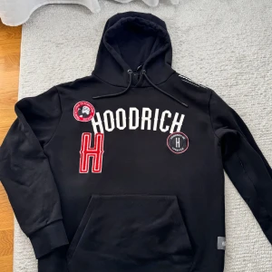 Svart hoodie från Hoodrich med patchar - Svart hoodie från Hoodrich med broderade patchar och stor vit logga framtill. Tröjan har huva med snörning, känguruficka och coola röda och vita detaljer. Tillverkad i 80% bomull och 20% polyester för en skön och mjuk känsla.