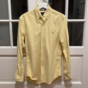 Gul skjorta från Ralph Lauren - Snygg ljusgul skjorta från Ralph Lauren i slim fit-modell. Klassisk design med knappar hela vägen, krage och broderad logga på bröstet. Tillverkad i mjuk bomull och har långa ärmar. Perfekt för dig som gillar stilrena och fräscha plagg.