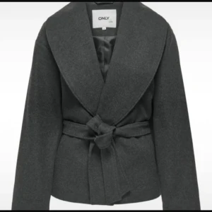 Mörkgrå kavajjacka från ONLY - Snygg mörkgrå kavajjacka från ONLY med bred krage och knytskärp i midjan. Jackan har lång ärm och en stilren, minimalistisk look som passar perfekt till lager-på-lager. Materialet är mjukt och känns lyxigt.