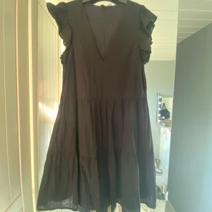 Svart volangklänning med v-ringning - Säljer en svart klänning med v-ringad hals och volangdetaljer på axlarna. Klänningen har en luftig, kort modell med flera lager och är ärmlös. Perfekt för dig som gillar en enkel men ändå cool stil❤️