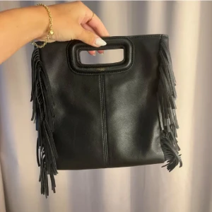 Svart axelväska med fransar från Maje - Säljer en svart axelväska från Maje i skinn med coola fransar på sidorna och ett stilrent handtag upptill. Väskan har en avtagbar axelrem och kommer med dustbag. Perfekt för dig som vill ha en unik och trendig accessoar. Köpt här på Plick, men kommer inte till användning.