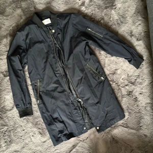 Mörkblå  lång bomberjacka med dragkedjor - Säljer en mörkblå  lång bomberjacka från Elvine med coola dragkedjedetaljer på ärm och fickor. Jackan har ribbade muddar, hög krage och tvåvägsdragkedja framtill. Perfekt för dig som gillar streetstyle och vill ha en stilren look.