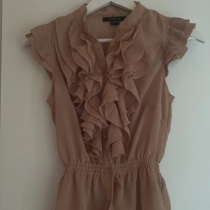 Beige kortklänning med volanger - Supersöt beige kortklänning med volangdetaljer framtill och knappar längs bröstet. Klänningen har ett smockat midjeparti som ger en snygg passform och är tillverkad i ett lätt, skirt material. Perfekt för dig som gillar romantisk och feminin stil.