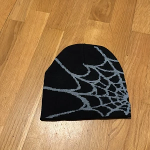 y2k - Cool svart mössa med grått spindelnätsmönster som täcker ena sidan. Perfekt accessoar för dig som vill sticka ut och addera lite edge till din stil. Tillverkad i stickat material som håller dig varm under kyliga dagar.