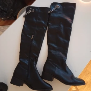 Svarta overknee boots med kedjedetalj - Snygga svarta overknee boots i syntetmaterial med silverfärgad kedja upptill och dragkedja på sidan. Skorna har låg klack, spetsig tå och platt sula. Perfekta för dig som vill sticka ut med en edgy look.