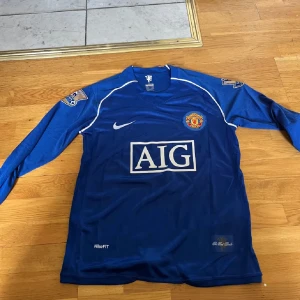 Manchester United Ronaldo 7 blå långärmad - Blå långärmad Manchester United fotbollströja från Nike med AIG som sponsor, klubbmärke på bröstet och Ronaldo 7 tryckt på ryggen. Tröjan har vita detaljer och Premier League-patch på ärmarna. Tillverkad i lätt och ventilerande material.