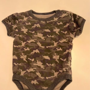 Camo kortärmad body i bomull - Kortärmad body med grå och gröna camouflage-mönster. Mjukt bomullstyg och praktiska tryckknappar vid axeln för enkel påklädning. Perfekt för en chill och cool look till de minsta.