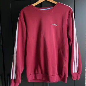 Vinröd Adidas sweatshirt med ränder - Vinröd sweatshirt från Adidas med klassiska vita ränder längs ärmarna och liten Adidas-logga på bröstet. Tröjan har rund halsringning, ribbade muddar och är långärmad. Perfekt för en sportig och avslappnad stil.